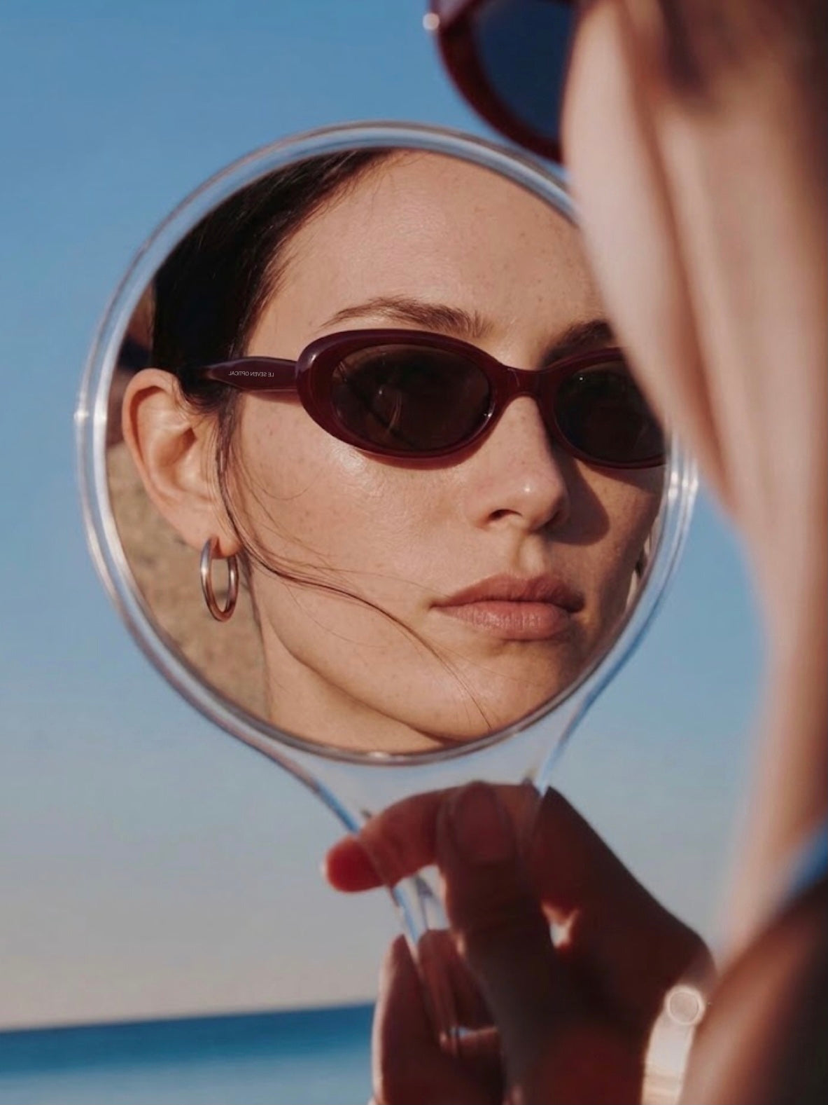 Woman wearing burgundy Sedona sunglasses reflected in a handheld mirror