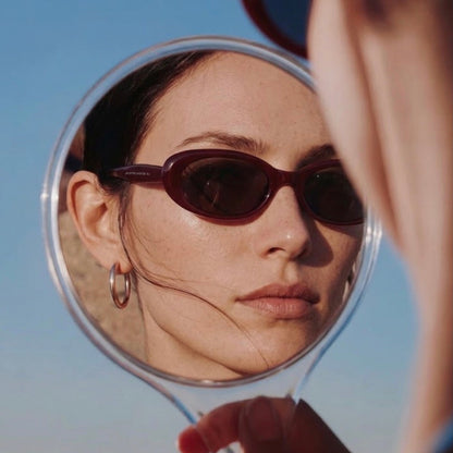 Model viewing Sedona Burgundy oval sunglasses in a hand mirror